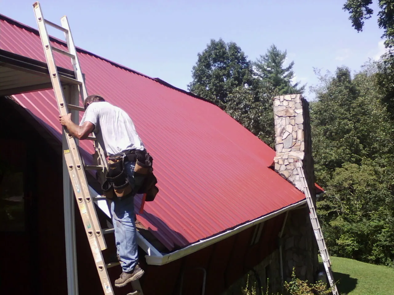Roofer on a ladder beside a red metal roof and stone chimney — Soffit & Fascia Repair by Roofing Brothers in Benton Harbor
